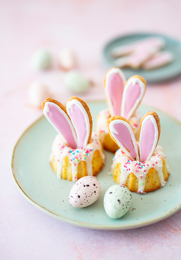 Kleine Gugelhüpfchen mit Hasenohren, dekoriert mit Puderzucker auf einem weißen Teller.